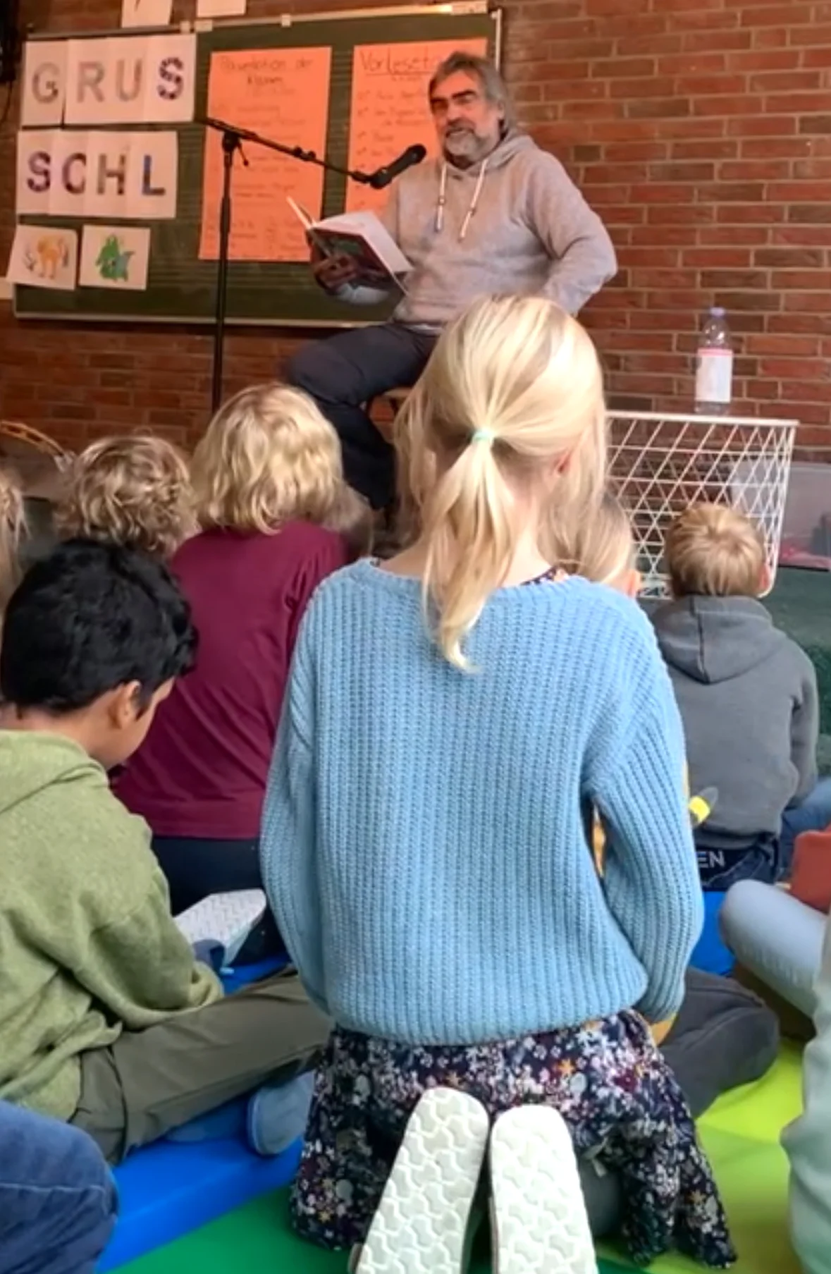 Ein Mann liest Kindern aus einem Buch vor, während diese auf bunten Matten vor ihm sitzen; im Hintergrund hängen Plakate und Buchstaben an einer Tafel. Ein Mann liest Kindern aus einem Buch vor, während diese auf bunten Matten vor ihm sitzen; im Hintergrund hängen Plakate und Buchstaben an einer Tafel.