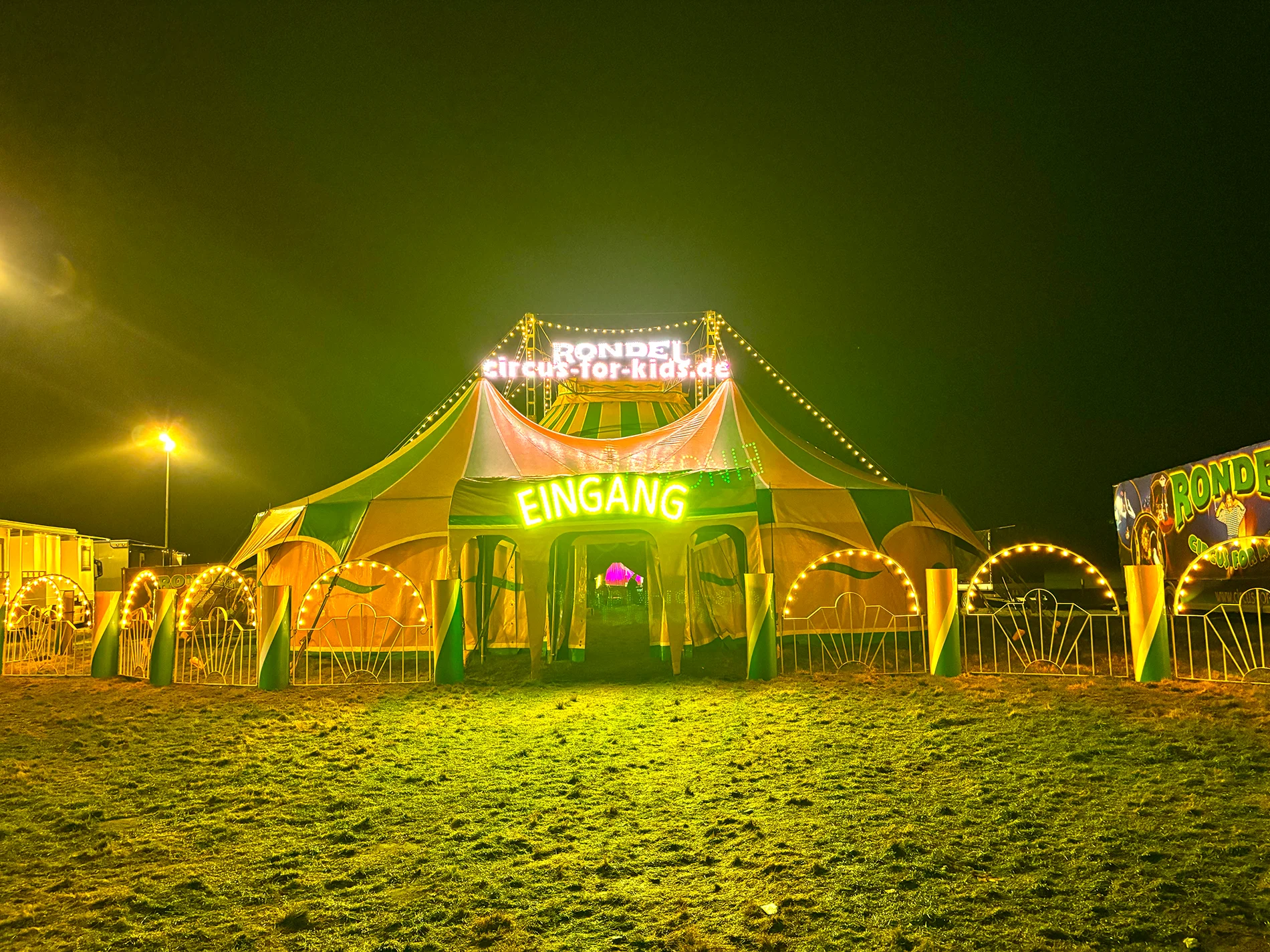 Zirkuszelt bei Nacht mit bunten Lichtern und großem „Eingang“-Schild. Zirkuszelt bei Nacht mit bunten Lichtern und großem „Eingang“-Schild.