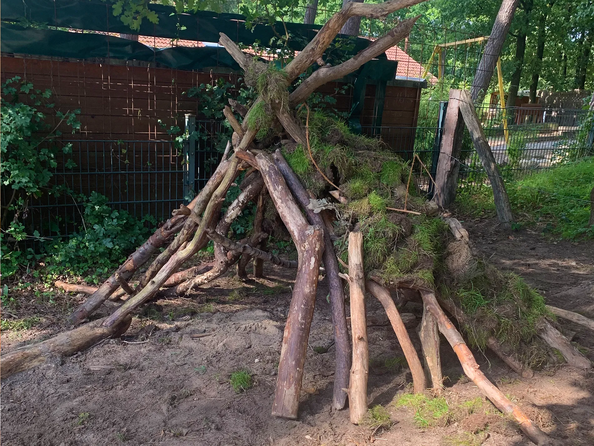 Ein aus Ästen und Gras gebautes Waldlager steht auf einem sandigen Boden im Außengelände der Schule. Das Spielhaus wurde kreativ aus Naturmaterialien errichtet und steht im Schatten von Bäumen. Ein aus Ästen und Gras gebautes Waldlager steht auf einem sandigen Boden im Außengelände der Schule. Das Spielhaus wurde kreativ aus Naturmaterialien errichtet und steht im Schatten von Bäumen.