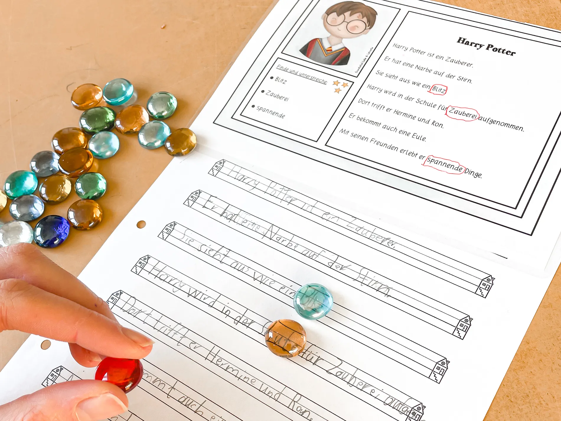 Ein Kind legt bunte Glassteine auf ein Arbeitsblatt zum Thema Harry Potter. Oben liegt ein Leseblatt mit Text und Illustration, unten ein Schreibblatt mit Linien und geschriebenen Sätzen. Ein Kind legt bunte Glassteine auf ein Arbeitsblatt zum Thema Harry Potter. Oben liegt ein Leseblatt mit Text und Illustration, unten ein Schreibblatt mit Linien und geschriebenen Sätzen.
